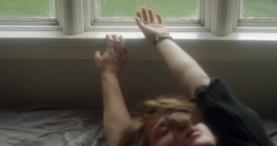 A woman lies in bed and reaches towards a window
