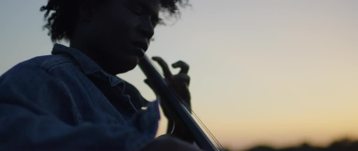 A woman plays the cello by the water at sunset with her eyes closed