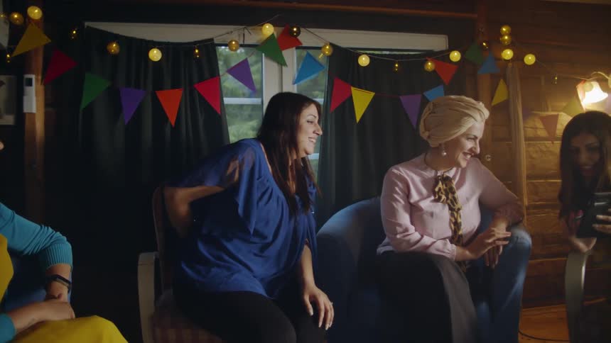 A woman walks into a party hiding a gift behind her back and surprising another woman with it