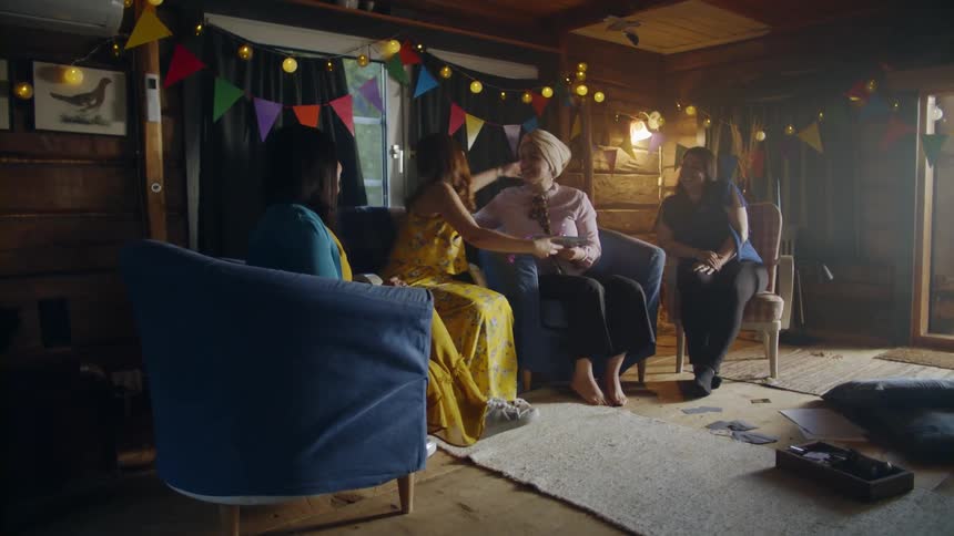 A group of woman sit in a room at a party and one hands a gift to another and hugs her
