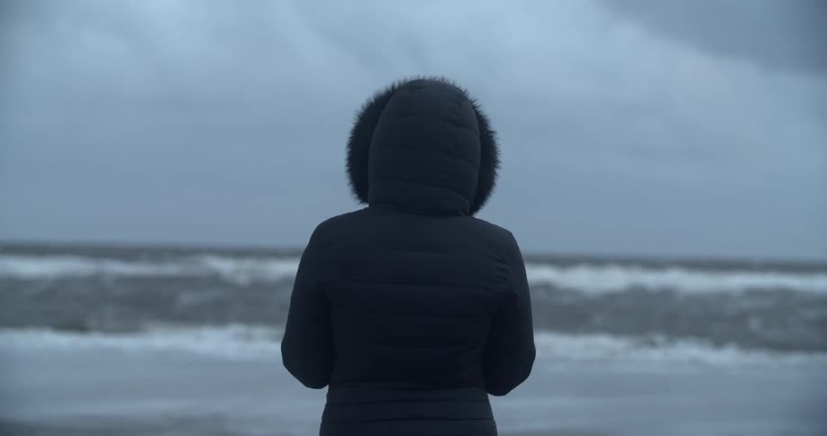 Person wearing hooded jacket it standing and facing ocean on cloudy day