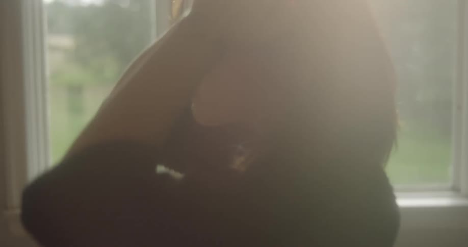 A woman walks around in front of a window on a sunny day and plays with her hair