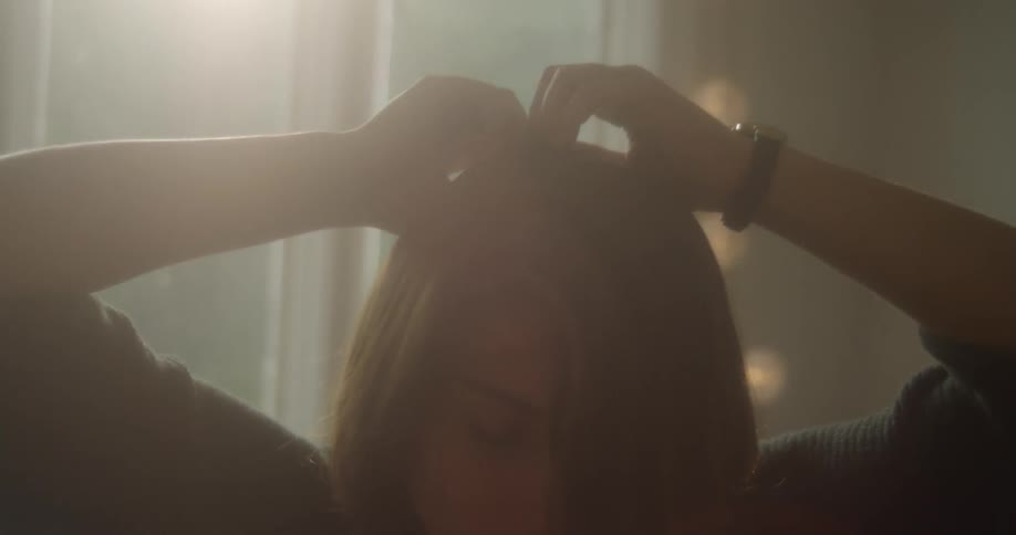 A woman stands in front of a window touching her hair with both hands