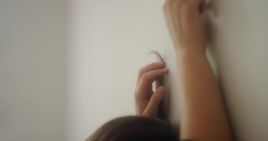 A woman stands with her head against a wall reaching up