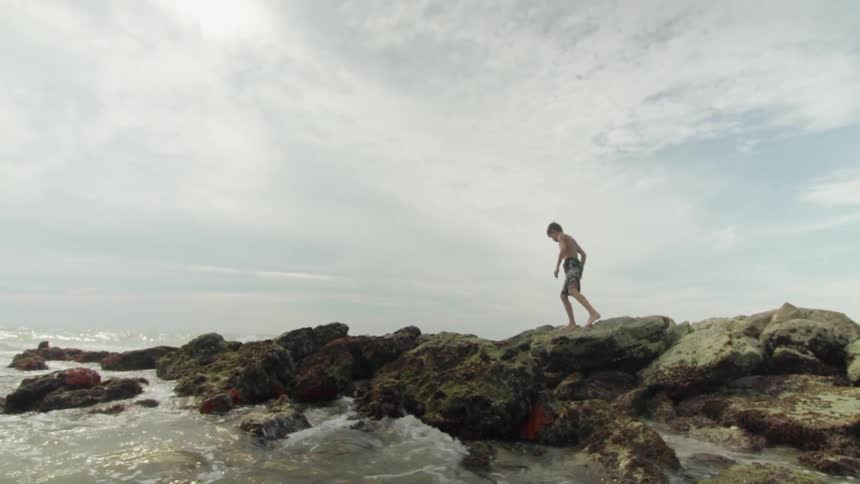 A boy carefully walks on the rocks as ocean waves splash against them