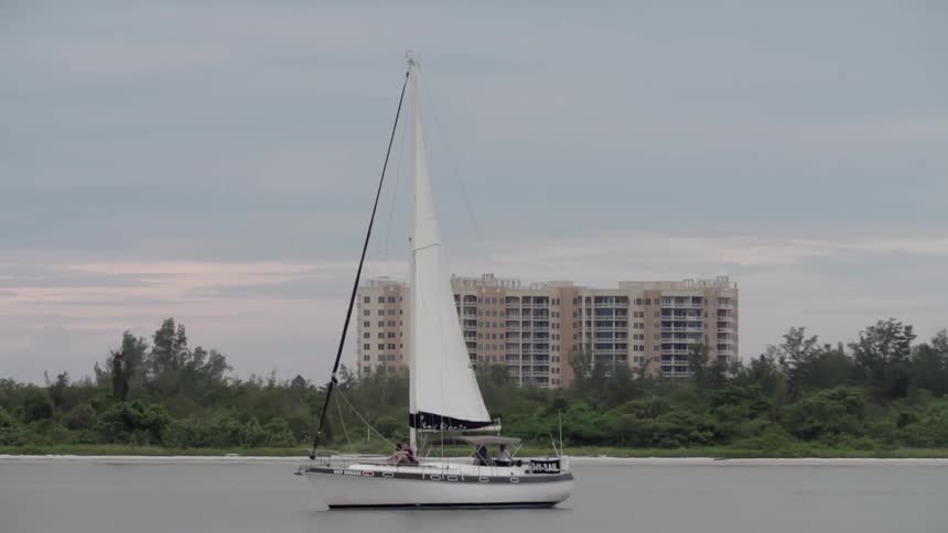 A sailboat moves across water with a building in the background on a cloudy day