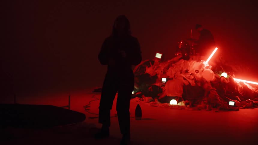 Person kneels on the floor while a man drums on a set next to a pile of objects and flashing lights