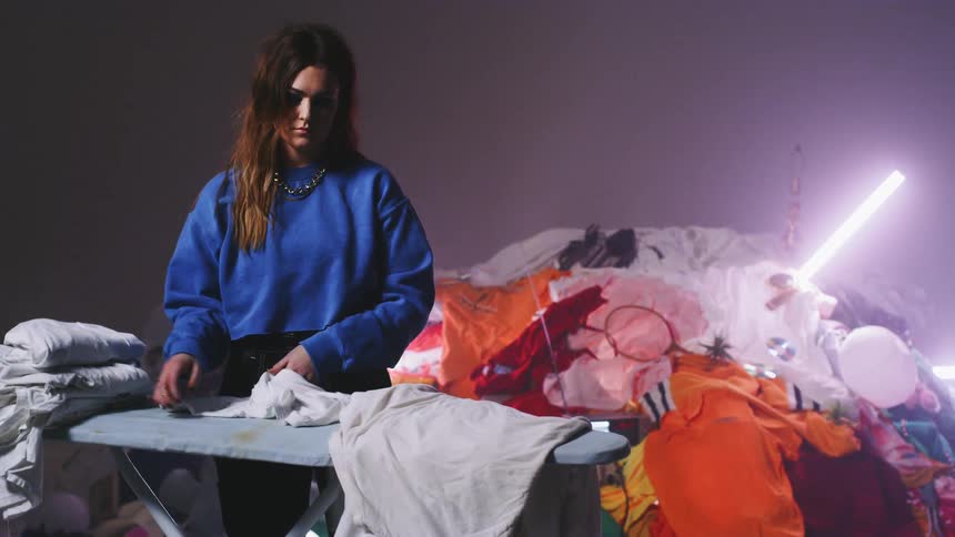 A woman folds laundry in front of a pile of clothes and random objects