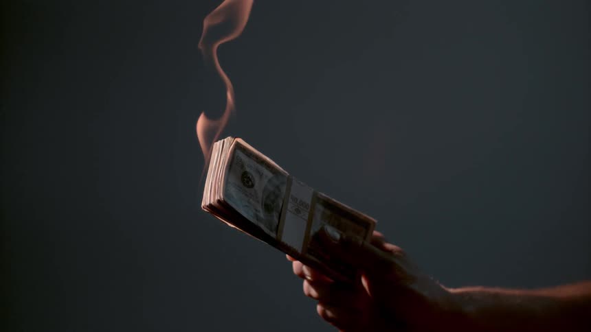 A hand holds a flaming bundle of banded bills