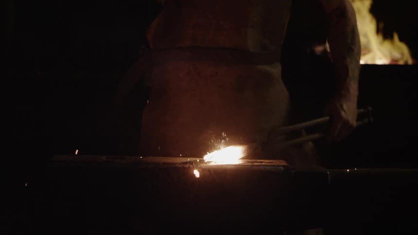 A blacksmith hammers metal in the dark in front of a fire