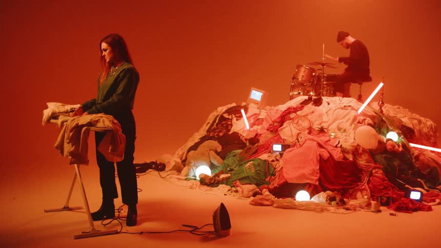 A folds laundry as a man plays drums on a mound of clothes