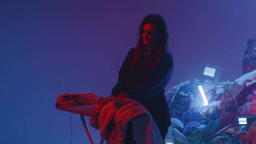 A woman folds laundry while a drummer plays on top of a pile of random objects
