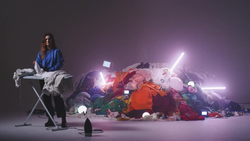 Woman folding clothes on an ironing board in front of a big pile of clothes in a studio