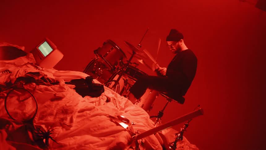 Drummer playing in a room next to a heap of various items with lights flashing in a reddish tint