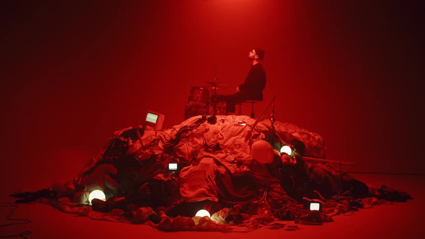 A  drummer sits on top of a pile of random objects