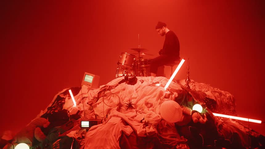 A drummer plays on top of a pile of random objects