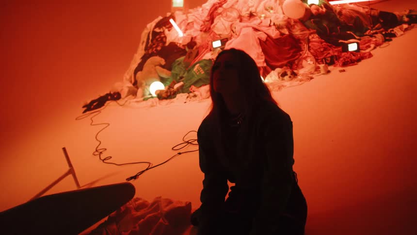 A woman sits in a dark room by a pile of clothes