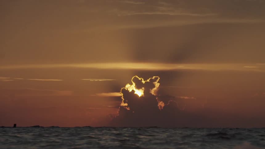 A view of the sun setting behind billowing clouds while choppy waves roll in the foreground