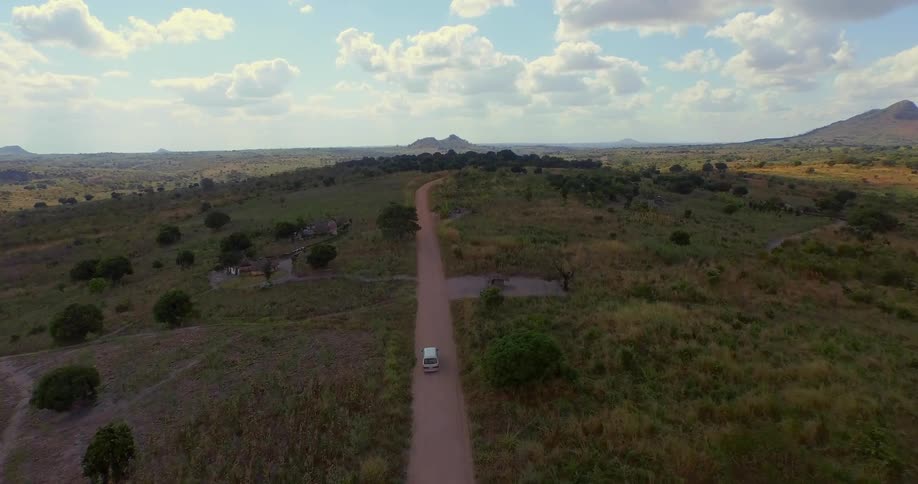 Aerial view of a vehicle driving down a small road with huge fields on the side and a cloudy sky