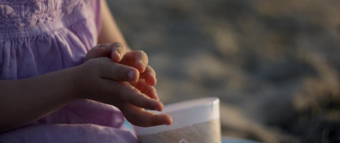 A toddler girl is sitting on a beach in daylight exploring the sand on her hands