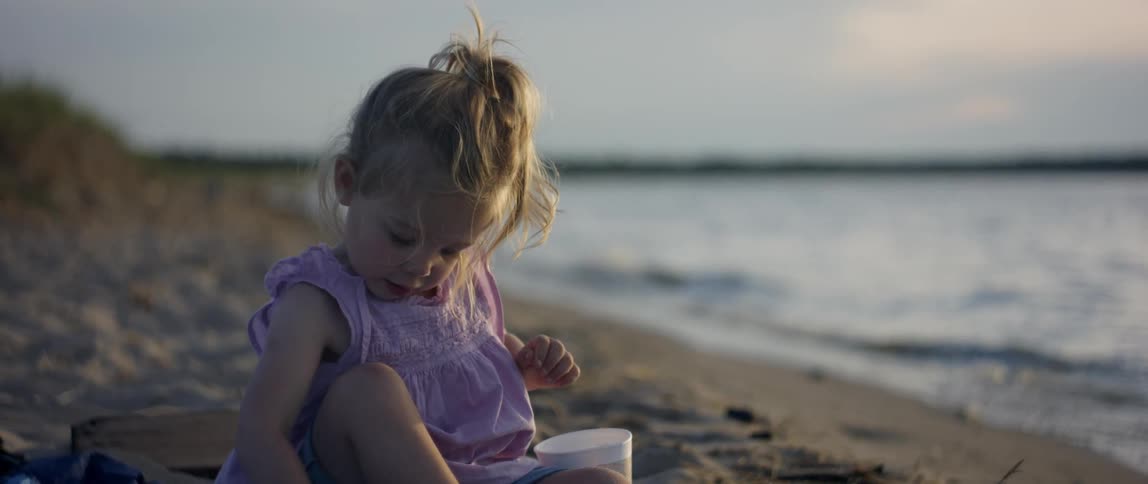 A toddler girl is playing in the sand at a beach in late afternoon