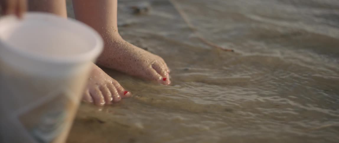 A wave laps the feet of a toddler girl as she stands on the beach in daylight