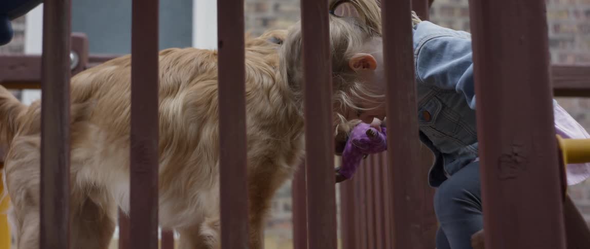 Little girl bends over while holding her doll as a dog nuzzles her