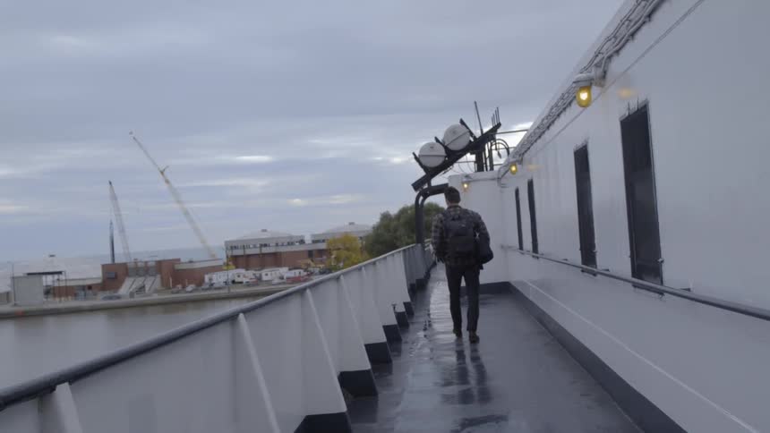 A man wearing a backpack walks on a wet walkway looking out at the water and up at the sky