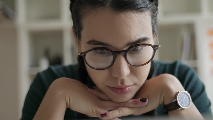 A woman wearing glasses is pausing to think and read while she sits working inside in daylight