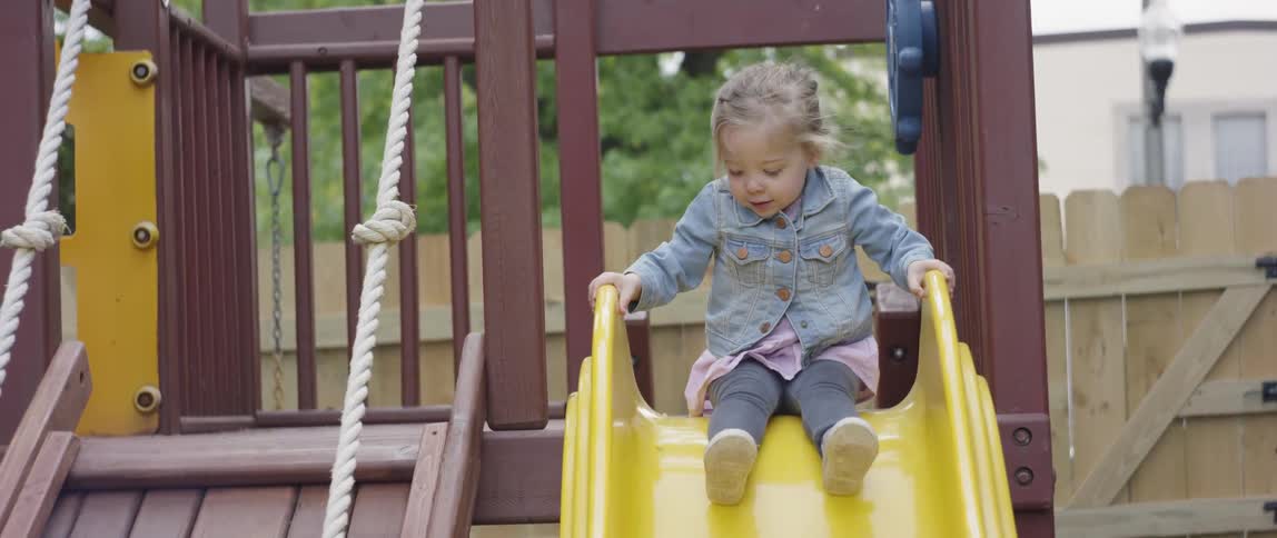A little girl is sitting down atop a slide on a backyard playground in daylight