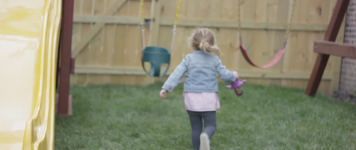 Little girl gets off of a slide while carrying her doll and runs towards the fence