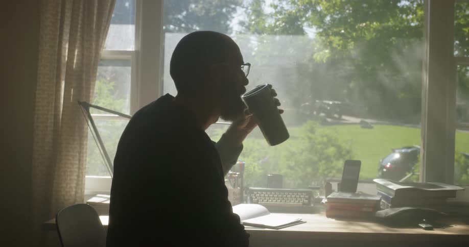 A man is taking a drink from a cup as he draws at a desk by a window in daylight