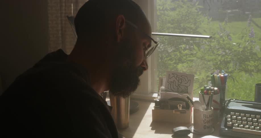 A man writes at a desk next to a sunny window