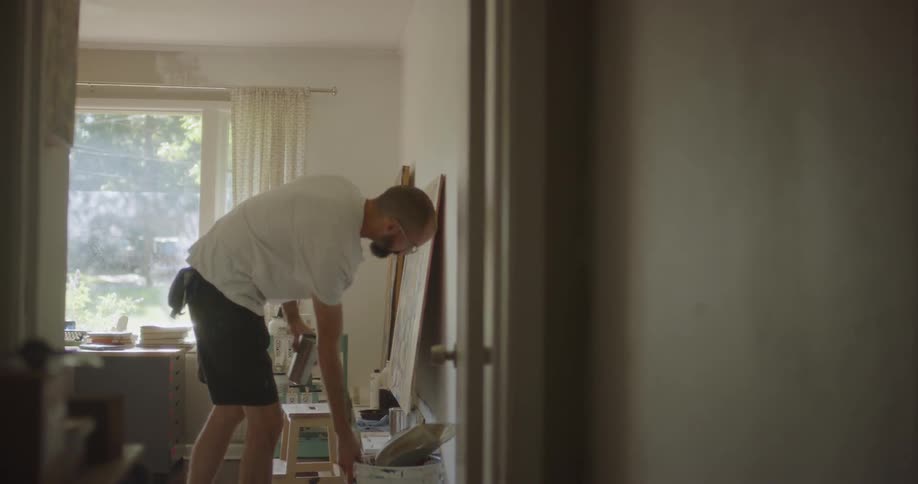 An artist is picking up his hat and a bucket and leaving his studio room inside in daylight