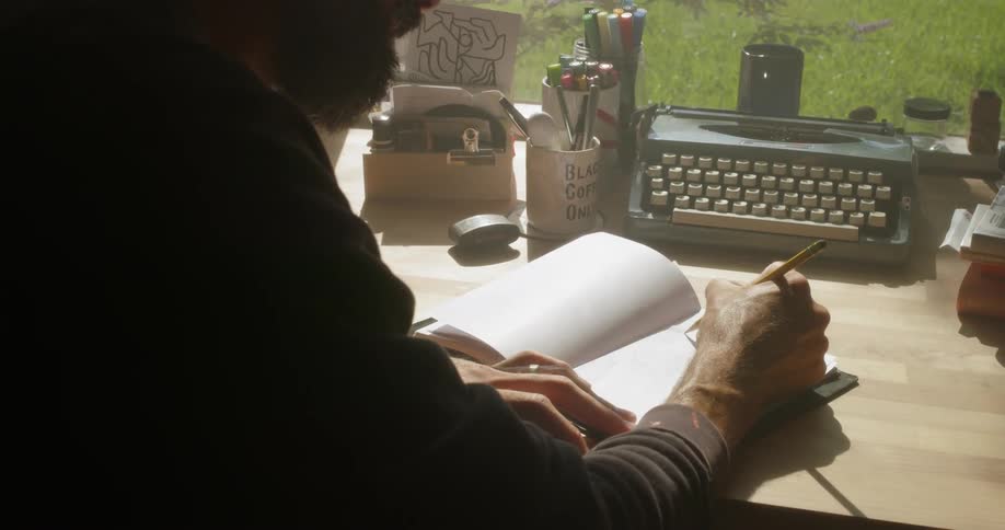 A man writes at a desk next to a sunny window