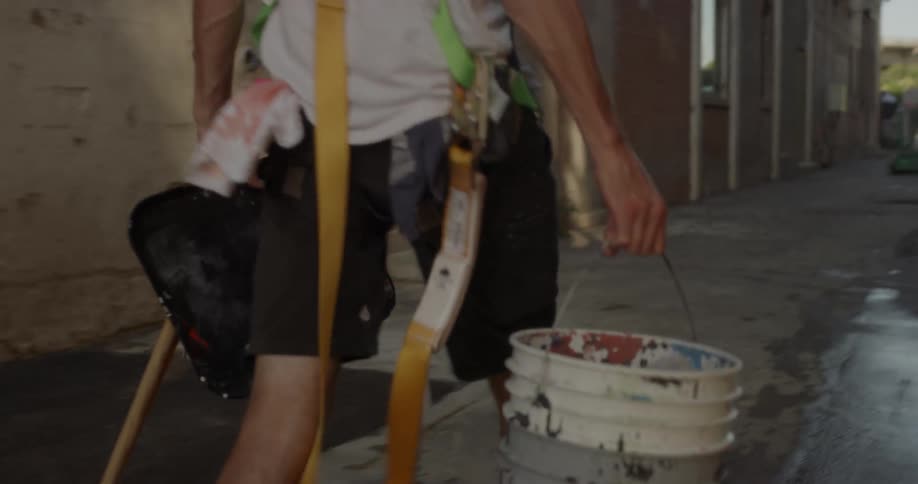 The man walks down the alley carrying a bucket and tools