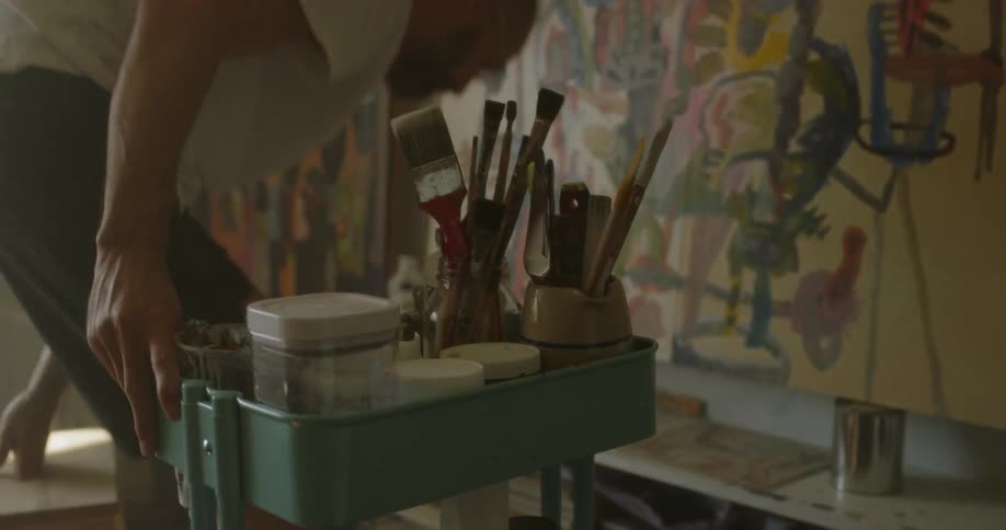 The man rolls a car full of brushes over to a painting