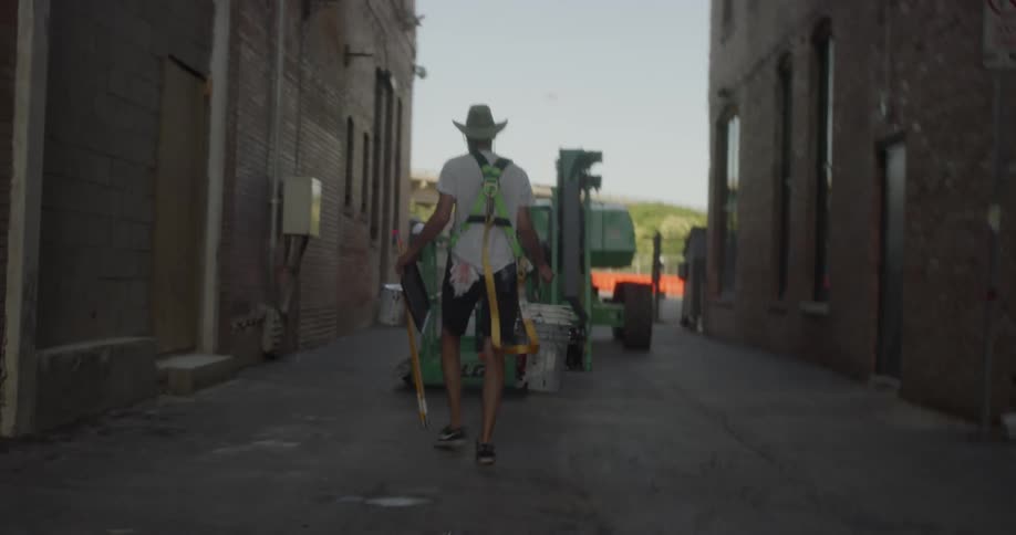 An artist is carrying a bucket and long paint brushes towards a crane in an alley in daylight