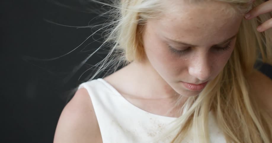 A blond teenage girl holds her hand to her head and looks down at something below camera
