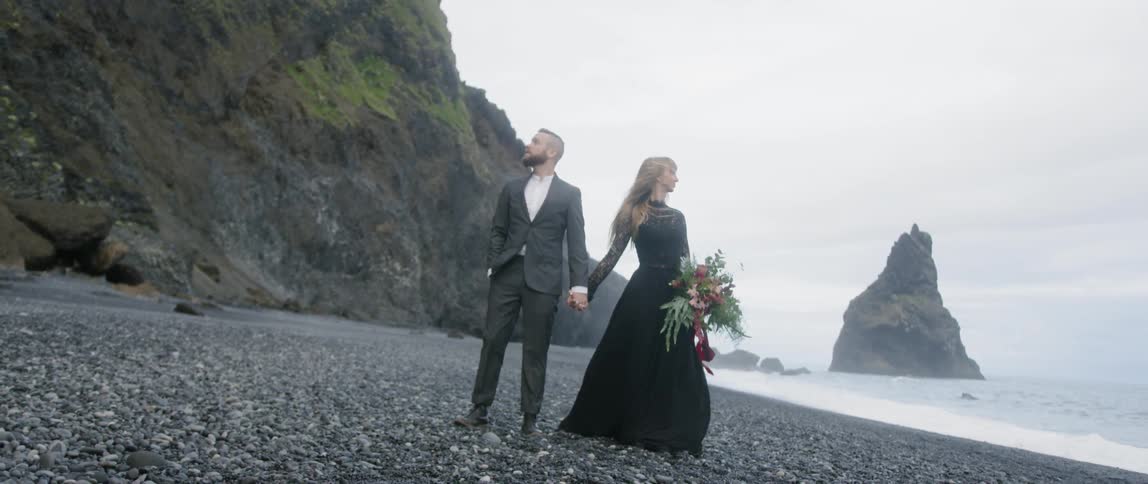A man and woman in suit and dress stand on the grey beach