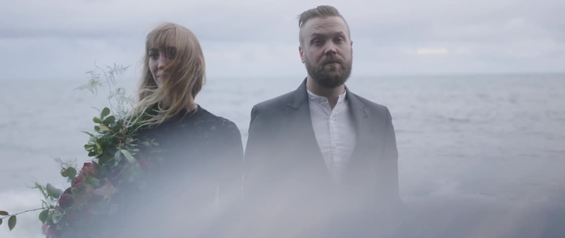 The two people stand together by the shore