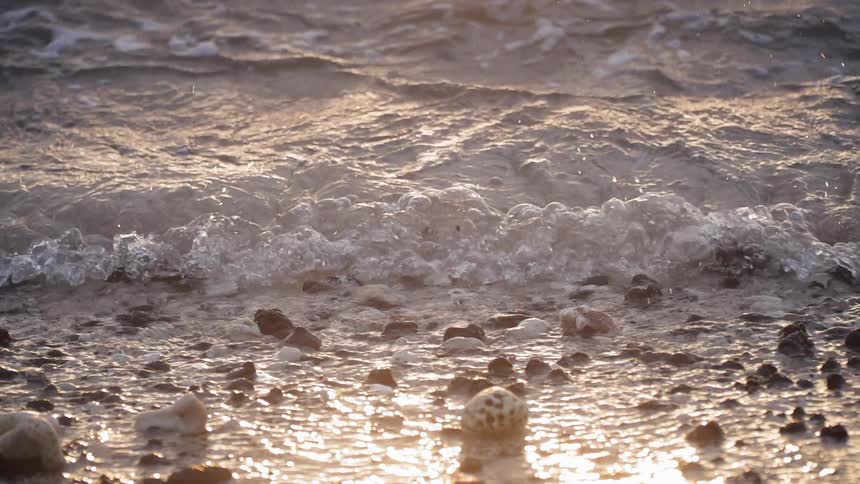 A waves is splashing and rolling over rocks on a beach at sunset