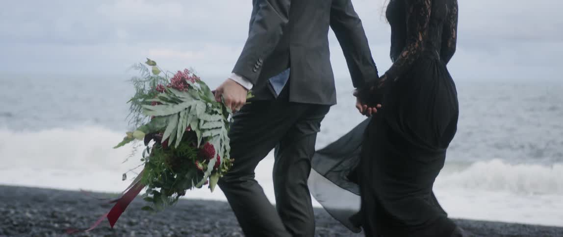 A man and woman hold hands while walking on a beach with a bouquet of flowers