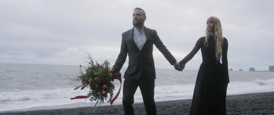 A man and woman hold hands on a beach with a bouquet of flowers