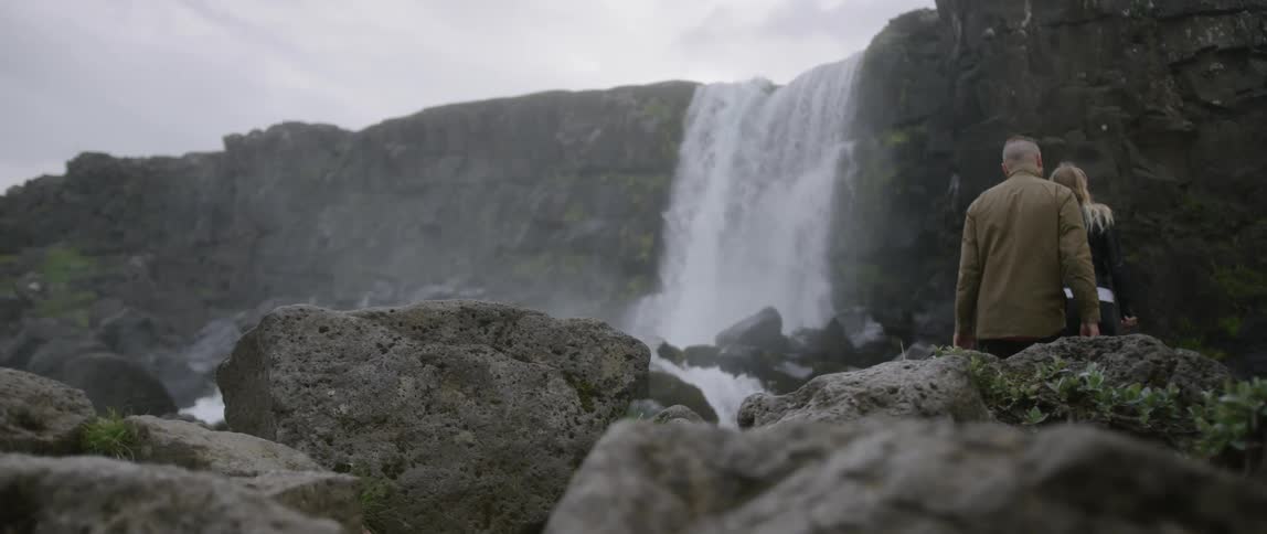 A man and woman walk towards a waterfall