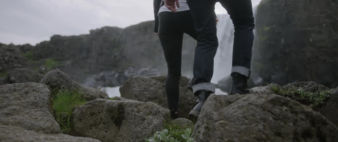 A man and woman walk over rocks near a waterfall