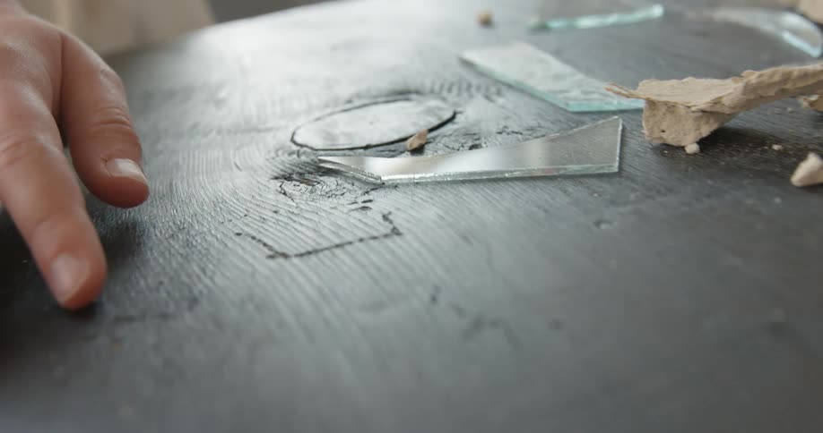 A hand drags across the surface of a table strewn with broken glass, ceramic and other materials