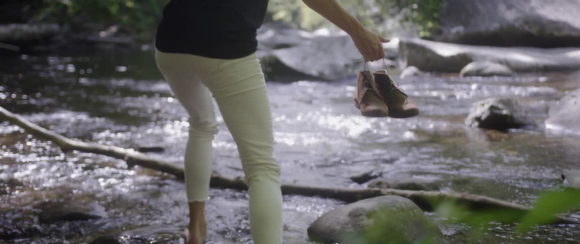 The woman holds her shoes as she walks in the stream