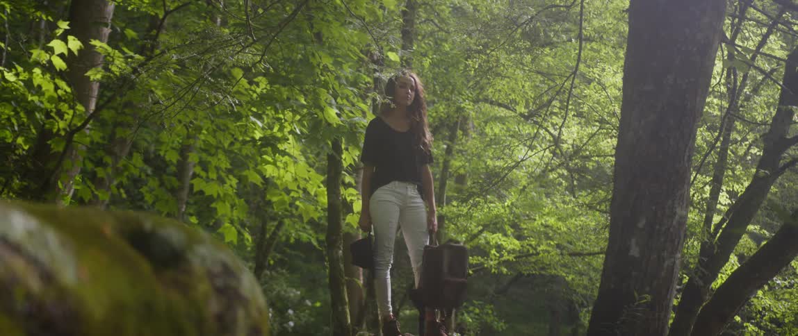 A woman stands holding a hat and bag in a forest