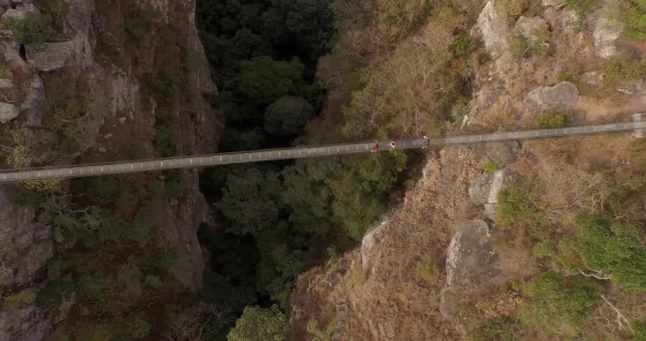 Overhead aerial view of three people crossing a narrow walking bridge over a deep chasm